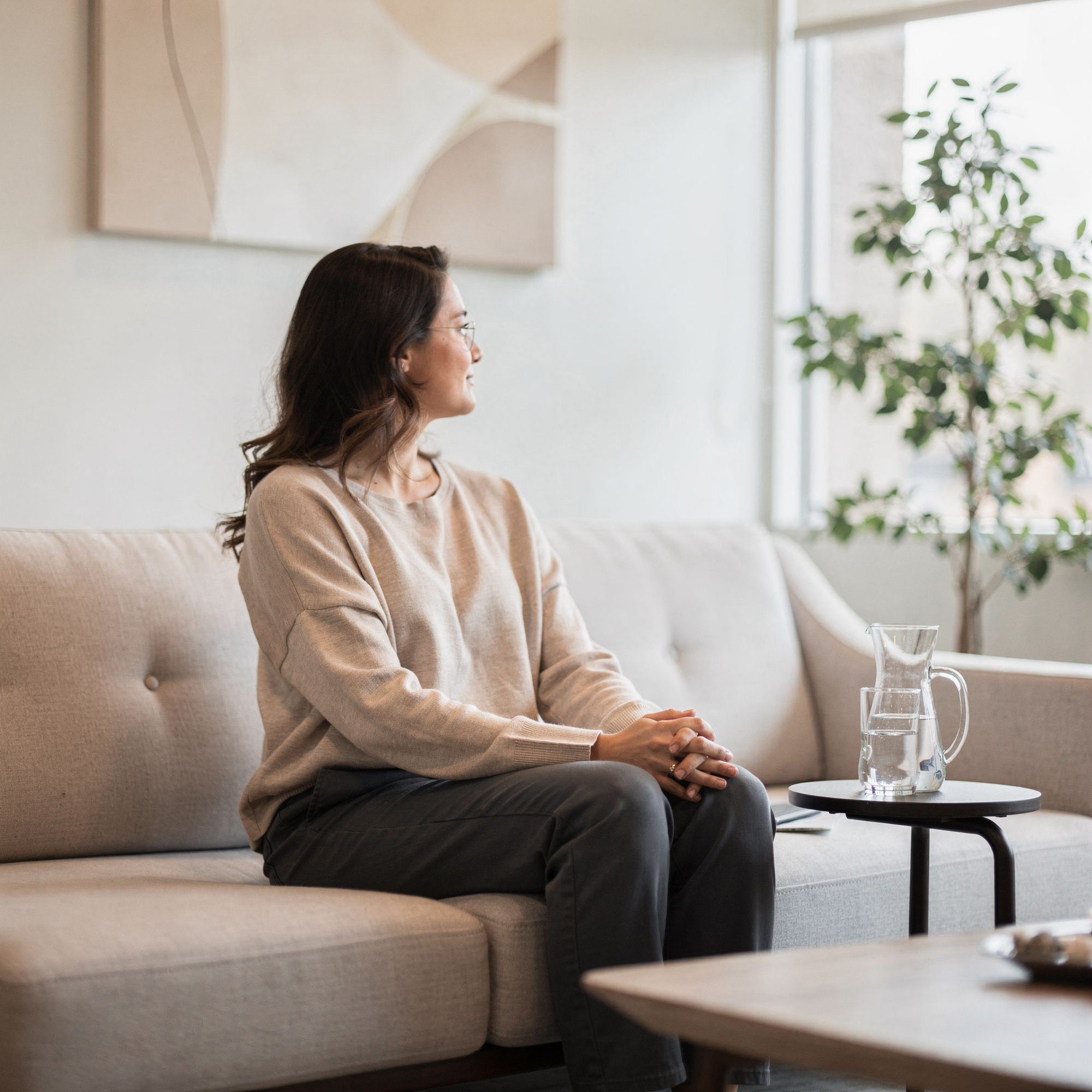 Woman sitting on a couch in a bright, modern clinical office with plants and water pitcher on side table